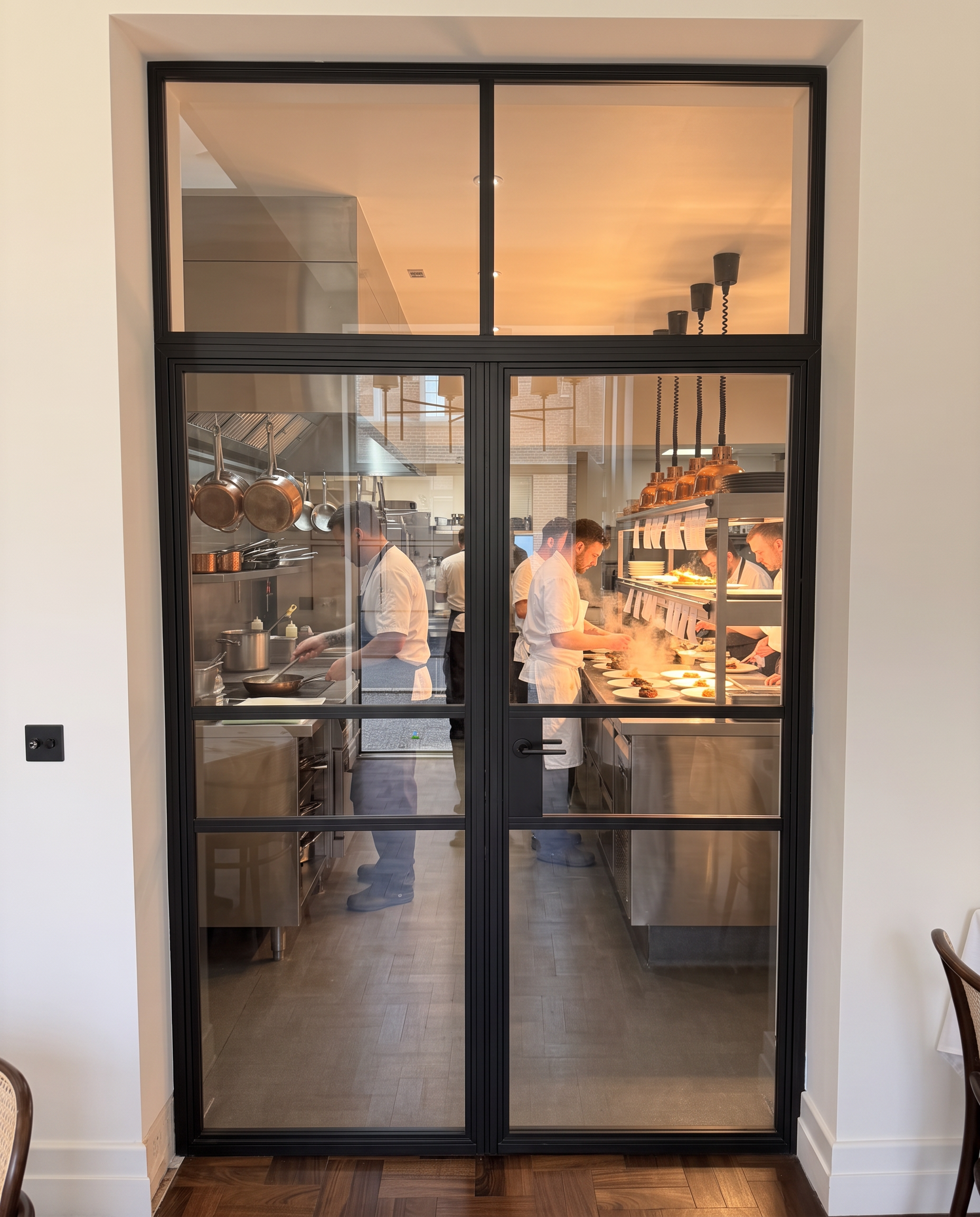 Matt black aluminium glazed screen and doors separating a restaurant dining room from a busy commercial kitchen